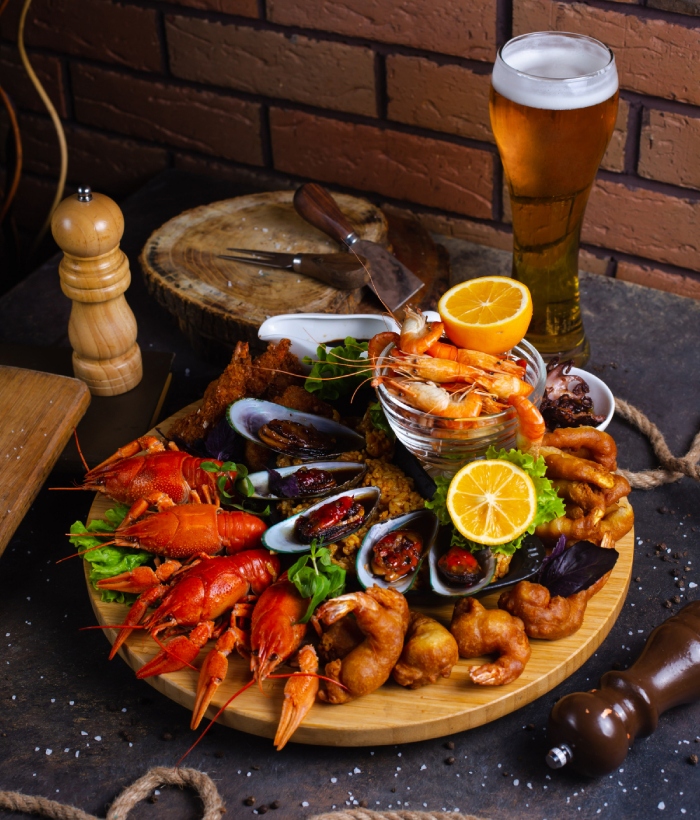 Plateau gourmand de fruits de mer avec homards, crevettes, moules, citron et bière, présenté sur une planche en bois.